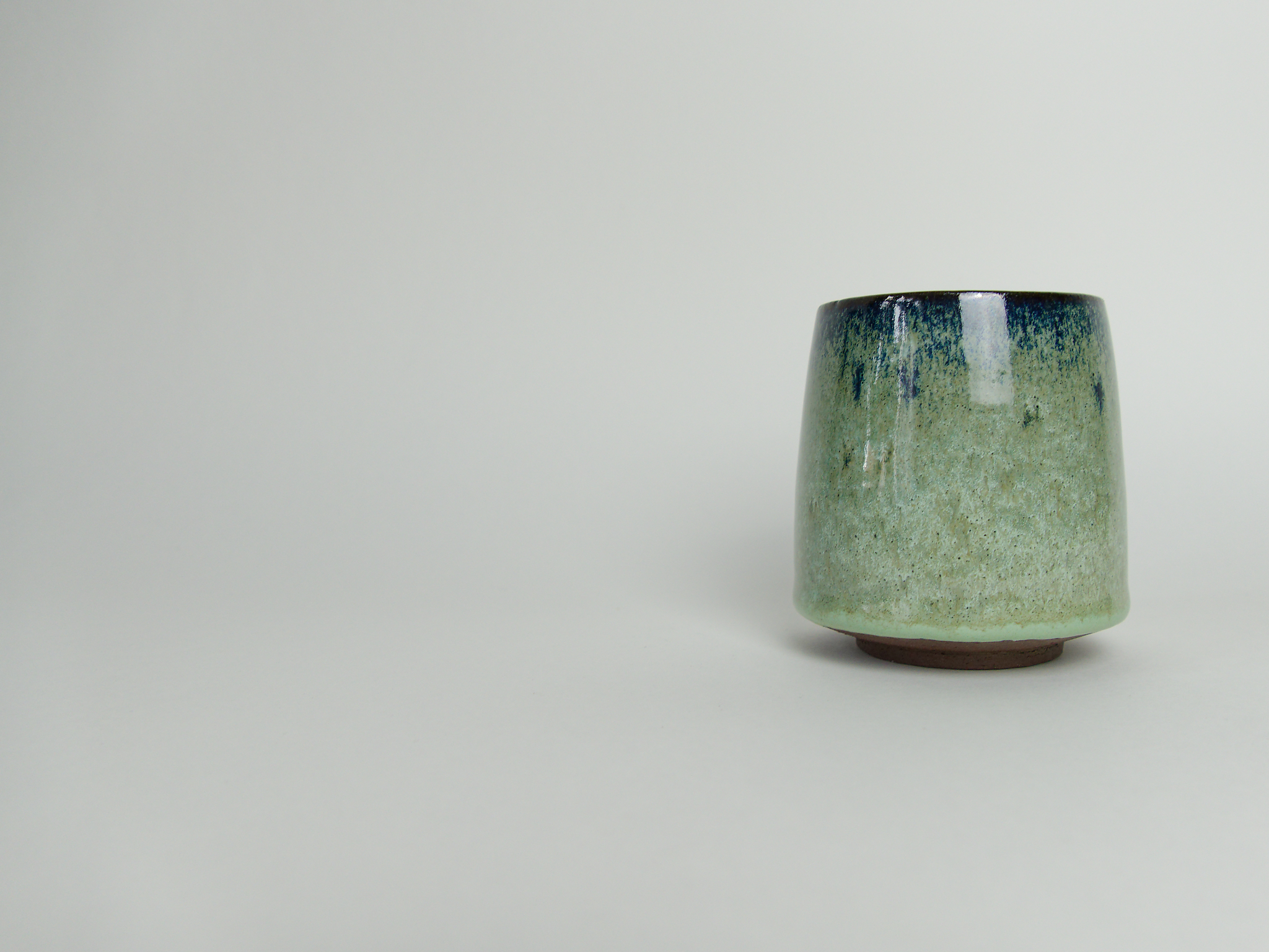 Handmade stoneware cup with a pale green glaze and dark blue rim on a light background.